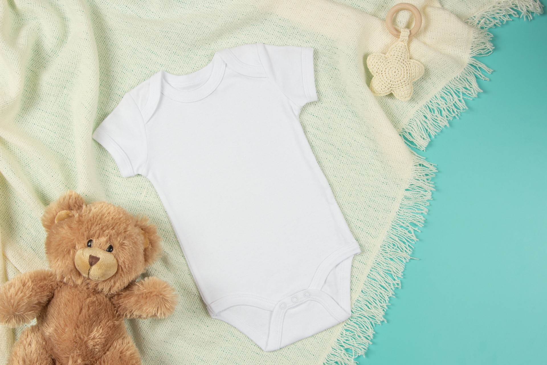 White cotton baby short sleeve bodysuit, teddy bear and natural wooden toy on beige blanket throw background. Infant onesie mockup. Blank gender neutral newborn bodysuit mock up template. Top view