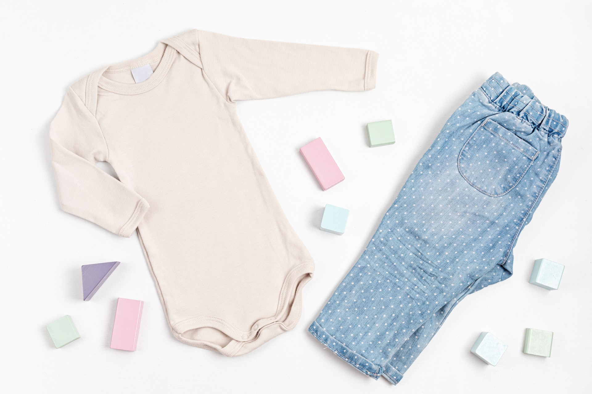 Composition with baby beige bodysuit, jeans, and toy building blocks on white background. Gender neutral child clothes